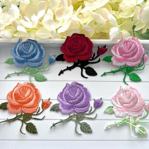 May include: Set of 8 embroidered iron-on rose patches in various colors, including pink, blue, purple, red, and orange. The roses are detailed with stitching and have green stems and leaves.