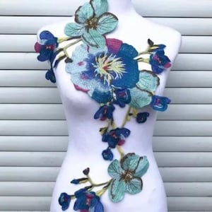 May include: A blue and green floral applique featuring a large blue flower in the centre. The applique is made of embroidered fabric and is ideal for adding a touch of elegance to clothing or crafts.