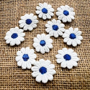 May include: A collection of ten white daisy flower patches with blue centers. The embroidered patches are arranged on a textured, brown burlap surface. Each flower has detailed petal stitching.