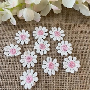 May include: A group of white daisy-shaped lace appliques with pink centres. The appliques are arranged on a textured, tan surface, with white and pink floral accents in the background. These delicate floral embellishments are ideal for craft projects.