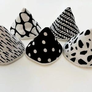 May include: Five cone-shaped fabric covers with a white fuzzy trim at the base. Each cover features a distinct black and white pattern, including irregular spots, polka dots, diagonal stripes, and organic shapes, arranged in a semi-circle formation.