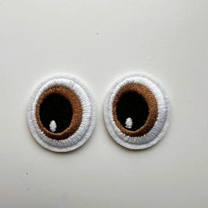May include: A pair of embroidered eye patches. Each patch has a brown iris, a black pupil, and a white highlight. The eyes are framed by a white, stitched border. These patches are suitable for crafts and adding a playful element to projects.