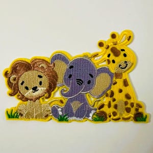 May include: Embroidered patch depicting a lion, elephant, and giraffe. The lion has a tan mane, the elephant is purple, and the giraffe has brown spots. All three animals are on a yellow background with green grass.