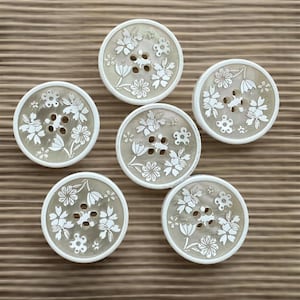 May include: Six round buttons with a translucent, light beige base and a white floral pattern printed on the surface. Each button features four central holes for attachment and a smooth, raised white border around the circular edge.