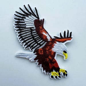 Könnte beinhalten: Ein gestickter Aufnäher, der einen Weißkopfseeadler im Flug mit weit ausgebreiteten Flügeln zeigt. Der Adler hat einen weißen Kopf und Schwanz, braunes Körpergefieder, schwarze Flügelspitzen sowie einen leuchtend gelben Schnabel und Krallen.