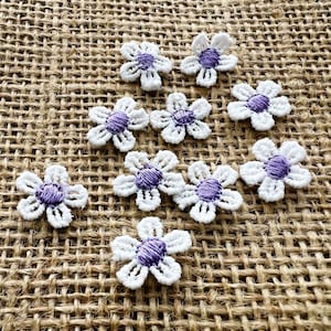 Könnte beinhalten: Zehn weiße Häkelblumen-Applikationen mit lila Zentren, angeordnet auf einer strukturierten, beigefarbenen Jutefläche. Jede Blume hat fünf Blütenblätter, ideal für Bastelprojekte.