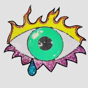 May include: A decorative eye-shaped patch with a green iris, white sclera, and a black pupil. The eye is surrounded by yellow flames, pink eyelashes, and a blue teardrop, all embellished with sequins.