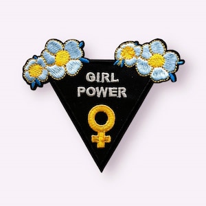 May include: Black triangular patch with the text "GIRL POWER" in white and a yellow female symbol. The patch is decorated with blue and yellow embroidered flowers.
