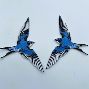 Könnte beinhalten: Zwei gestickte Schwalben-Aufnäher mit leuchtend blauem Gefieder, schwarzen Flügeldetails und weißen Akzenten. Jeder Vogel ist in einer dynamischen Flugpose mit ausgebreiteten Flügeln dargestellt, mit feinen Stickereien und einem kleinen rosa Punkt am Kopf.