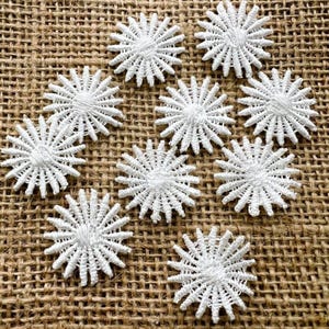 May include: Nine white, star-shaped lace appliques are arranged on a textured, brown burlap surface. Each applique features a central, circular design with radiating, pointed edges, creating a delicate, floral appearance. These appliques are ideal for craft projects.