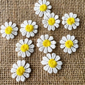 May include: Ten white fabric daisy flowers with yellow centres. The flowers are arranged on a brown hessian background.