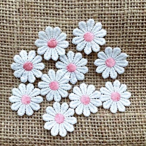 Könnte beinhalten: Neun weiße Gänseblümchen-Blumenaufnäher mit rosa Zentren sind auf einer strukturierten, braunen Jutefläche angeordnet. Die gestickten Blumen haben detaillierte Blütenblätter und sind ideal zum Basteln oder Verzieren von Kleidung.