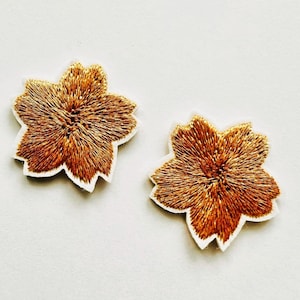 May include: Two flower-shaped patches featuring metallic gold embroidery thread in a dense, radiating stitch pattern. Each patch has a distinct five-petal silhouette with a clean white fabric border outlining the edges of the floral design.