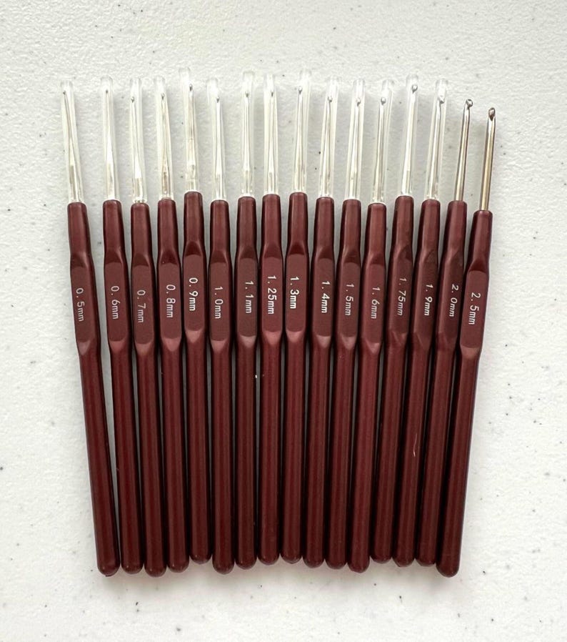 K&ouml;nnte beinhalten: Ein Set mit 16 H&auml;kelnadeln in Burgunderrot mit durchsichtigen Kunststoffgriffen. Die Nadeln sind nach Gr&ouml;&szlig;e sortiert, von 0,5 mm bis 2,5 mm. Jede Nadel ist mit ihrer Gr&ouml;&szlig;e in Millimetern gekennzeichnet.
