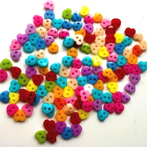 Peut inclure: Une collection de petits boutons en plastique en forme de cœur dans une variété de couleurs vives, notamment rouge, bleu, jaune, vert, rose, orange, violet et blanc. Chaque bouton comporte deux trous au centre pour la couture et une finition lisse.