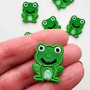 Könnte beinhalten: Gestickte grüne Froschaufnäher mit schwarzen Gesichtern und weißen Augen. Die Aufnäher sind klein und haben eine strukturierte Optik. Die Froschaufnäher sind ein lustiges Bastelzubehör.