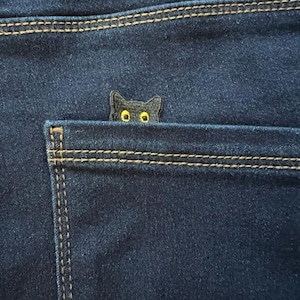 May include: Dark blue denim jeans with a small black cat patch peeking from the back pocket. The cat patch has yellow eyes and is stitched onto the pocket. The jeans have visible stitching.