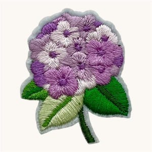 May include: A fabric patch featuring an embroidered hydrangea flower in shades of purple and white. The design includes detailed stitching for the petals and vibrant green leaves at the base, all set against a light gray border outline.
