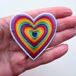 May include: Rainbow heart-shaped embroidered patch with a white outline. The patch is made of colourful thread and has a layered design.