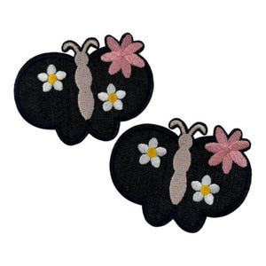 Könnte beinhalten: Zwei schmetterlingsförmige Stickaufnäher mit schwarzem Hintergrund in Denim-Optik. Jeder Aufnäher zeigt einen hellbeigen Körper, zwei weiße Blüten mit gelber Mitte und eine rosa Blüte mit fünf Blütenblättern, umrandet mit dunkelblauem Garn.