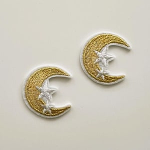 May include: Two embroidered patches featuring a crescent moon design with gold thread and white stars. The patches have a white border and are set against a white background. These patches are suitable for clothing or accessories.
