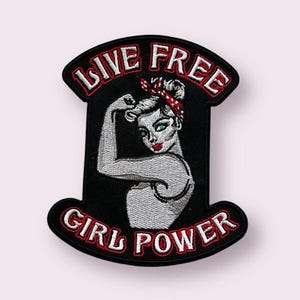 May include: A black embroidered patch featuring a stylized illustration of a person flexing their arm. The design includes a red and white polka dot headband, red lips, and the text "LIVE FREE" at the top and "GIRL POWER" at the bottom in white.