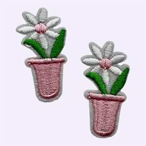 May include: Two embroidered iron-on patches, each featuring a white daisy with a pink centre, green leaves, and a pink flower pot. The patches have a light grey border and are designed with detailed stitching to create a textured floral appearance.