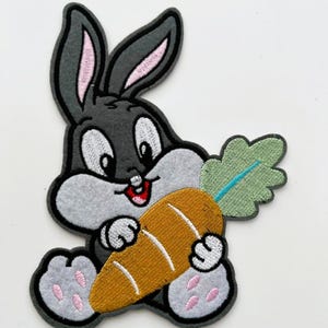 May include: Embroidered patch featuring a cartoon rabbit holding a carrot. The rabbit is gray with pink inner ears and paws. The carrot is orange with green leaves and a blue stem. The patch has a black border.