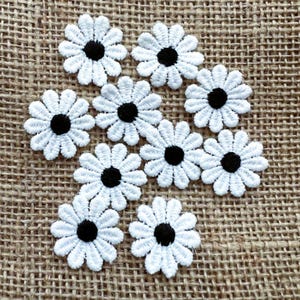 May include: A group of white daisy flower appliques with black centres. The appliques are made of delicate, embroidered fabric and are arranged on a textured, tan background. These floral embellishments are suitable for various craft projects.