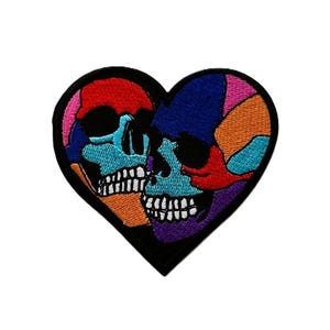 May include: A heart-shaped embroidered patch featuring two skulls in profile facing each other. The design uses vibrant, multi-coloured thread including red, blue, orange, purple, and teal, all outlined with a thick black border on a black background.
