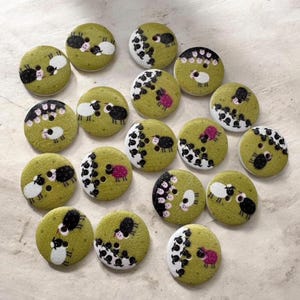 May include: A collection of round, olive green buttons, each featuring hand-painted illustrations of small sheep. The designs include various arrangements of white, black, and pink sheep figures, with some buttons showing single sheep and others displaying small groups.