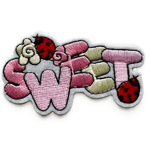 May include: Embroidered patch with the word "SWEET" in pink, with a black outline. The patch features a flower, ladybugs, and other floral elements in shades of pink, green, and white. The patch is on a gray background.