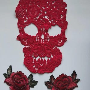 May include: A red lace skull applique with detailed patterns, alongside two embroidered red rose patches with green and black leaves. The skull and roses are on a white background.