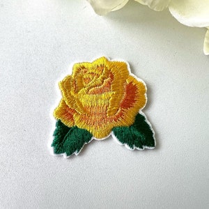 May include: An embroidered yellow rose with green leaves. The rose is a single bloom with a yellow centre and a layered design. The leaves are a dark green.