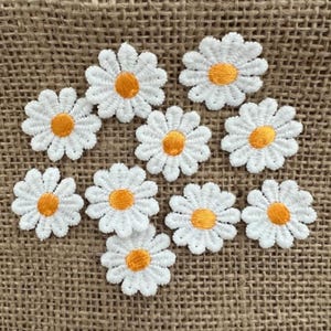 May include: Ten individual daisy flower patches featuring white embroidered petals and a bright yellow centre. Each patch is crafted with detailed stitching, showcasing a classic floral design with a circular shape and multiple rounded petals.