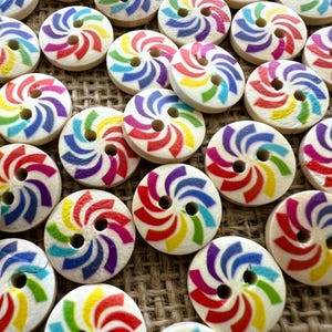 May include: A collection of round, two-hole wooden buttons, each featuring a vibrant, multi-colored spiral pattern in shades of red, orange, yellow, green, blue, and purple printed on a natural light-colored wood surface.