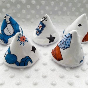 May include: Five cone-shaped fabric items featuring various printed patterns including blue cartoon characters, black stars, orange shapes, and blue geometric designs. Each item has a white base and a soft, white, textured fabric exterior.