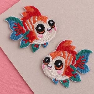 May include: Two embroidered iron-on patches shaped like goldfish. Each fish features a white face with large black eyes, pink cheeks, and a smiling mouth. The bodies are orange and white with vibrant red, blue, and green fins and tails.