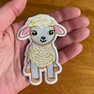 May include: A white and yellow embroidered patch of a cartoon sheep with pink ears. The sheep is standing on all four legs and has a friendly expression.