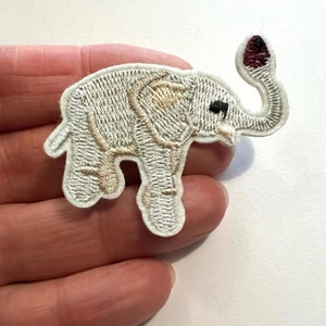 May include: White embroidered iron-on patch of an elephant with a raised trunk and a burgundy tip. The elephant has beige details on its body.