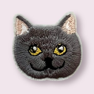 May include: Embroidered grey cat face patch with yellow eyes. The cat has a white patch on its ear and a small black nose.
