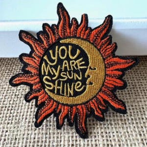 May include: Embroidered patch with a sun and crescent moon design. The sun is orange and red with black outlines, surrounding a tan crescent moon. The text "YOU ARE MY SUN SHINE" is embroidered in gold.