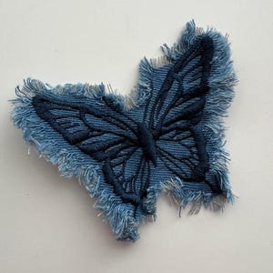May include: A denim butterfly patch with frayed edges. The butterfly design is embroidered in a darker shade of blue, creating a textured effect. The patch is likely for clothing or accessories.