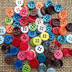 May include: A selection of multi-coloured round buttons, including shades of blue, green, red, and brown. Each button has four holes and is scattered on a textured, beige surface.