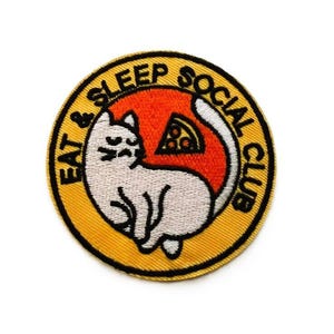 May include: A circular embroidered patch with a yellow border and black text reading EAT & SLEEP SOCIAL CLUB. The centre features a white cat curled into a circle against an orange background, with a small slice of pizza positioned above the cat.