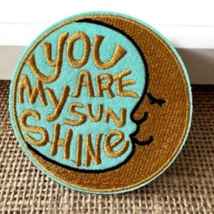 May include: Circular embroidered patch with the text "YOU ARE MY SUN SHINE" in gold thread. The design features a teal blue circle with the text and a crescent moon in gold. The patch has a brown border.