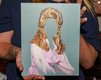 May include: A framed print of a woman with long, blonde hair styled in a large, voluminous bouffant. She is wearing a pink blouse with a white collar and a white lace trim. The background is a light blue color.