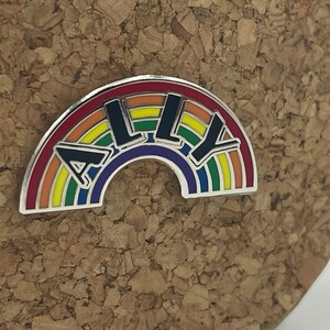 May include: A rainbow-colored enamel pin with the word "ALLY" in black block letters. The pin has a silver border and a rainbow design, with the colors red, orange, yellow, green, blue, and purple. The pin is on a cork surface.