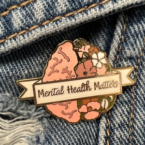 May include: A gold-colored enamel pin with a brain design, half of which is a light pink brain and the other half is flowers. A white banner across the bottom reads "Mental Health Matters".