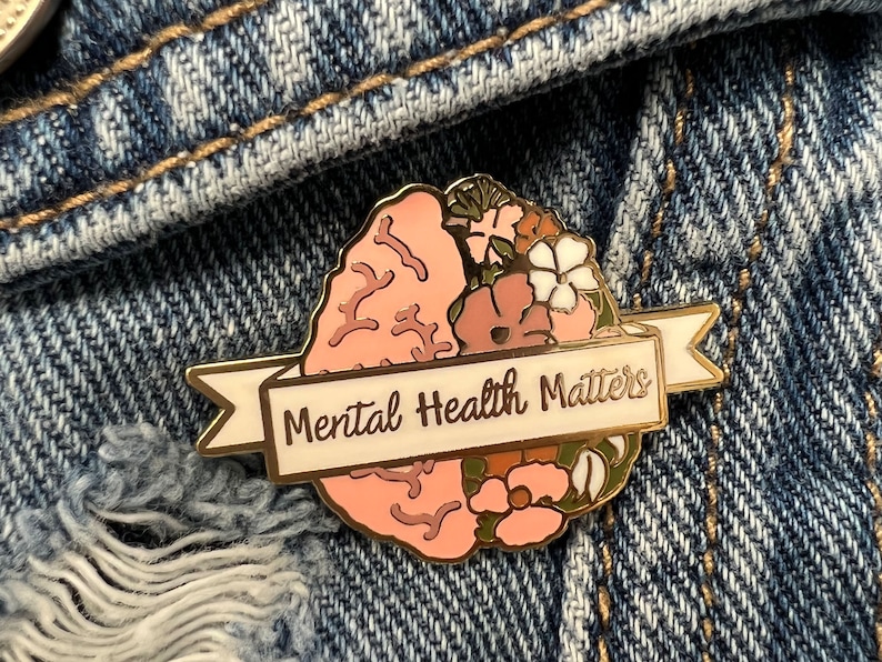 May include: A gold-colored enamel pin with a brain design, half of which is a light pink brain and the other half is flowers. A white banner across the bottom reads "Mental Health Matters".